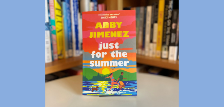 The book Just for the Summer by Abby Jimenez, cover facing front, in front of a library shelf